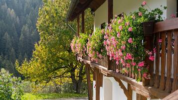 Un balcon en bois orné de fleurs roses, un bâtiment blanc et, en arrière-plan, une montagne couverte de forêts.