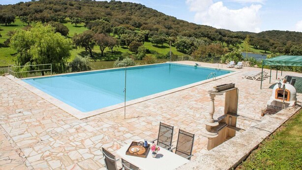 A rectangular swimming pool with a stone deck, surrounded by lounge chairs and a table with a tray of items.