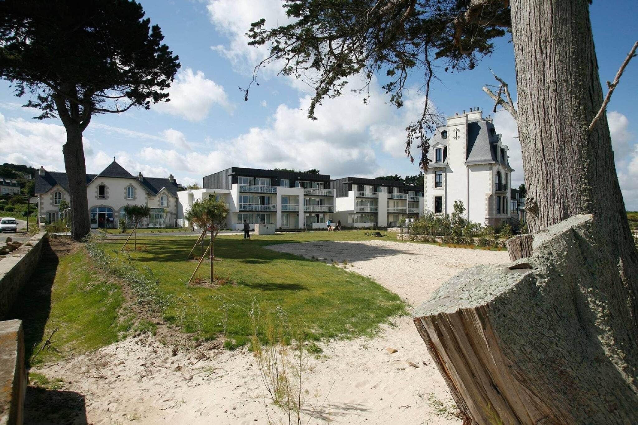 Un quartier résidentiel où se côtoient des bâtiments modernes et traditionnels, un sentier sablonneux et un grand arbre.