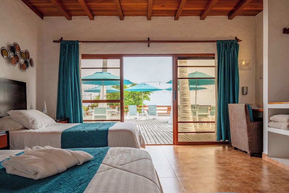 A hotel room with two beds, a wooden ceiling, a balcony with outdoor seating, and a view of the outdoors.