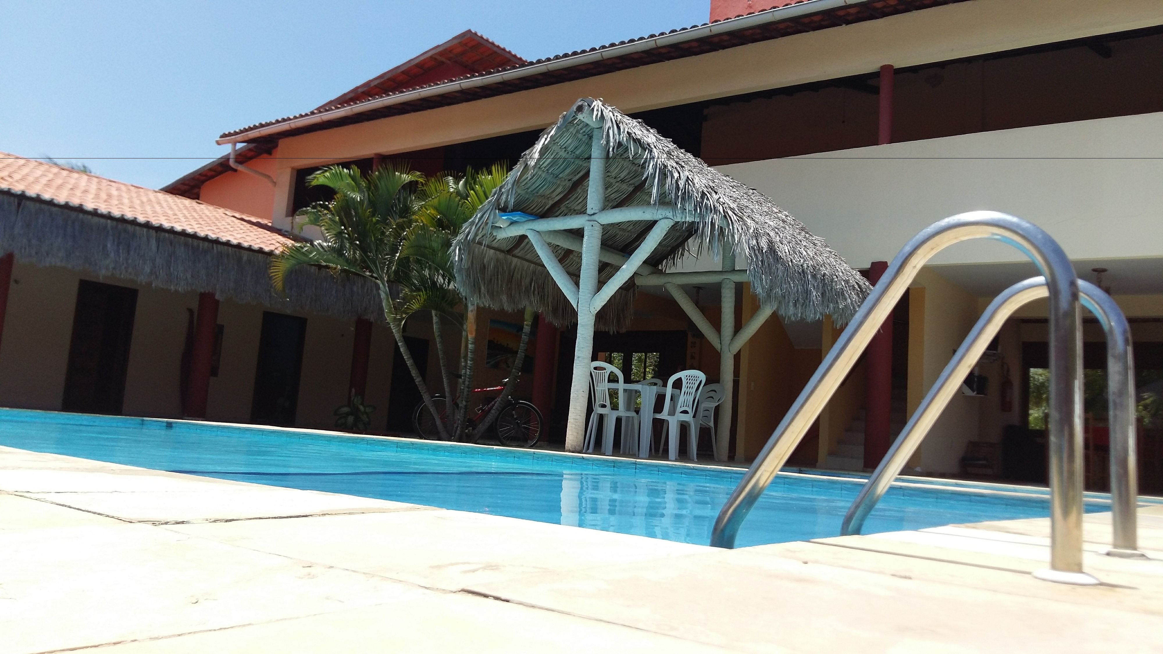 Une piscine dotée d’une structure de toit en chaume, de chaises en plastique blanc et d’une échelle en métal.