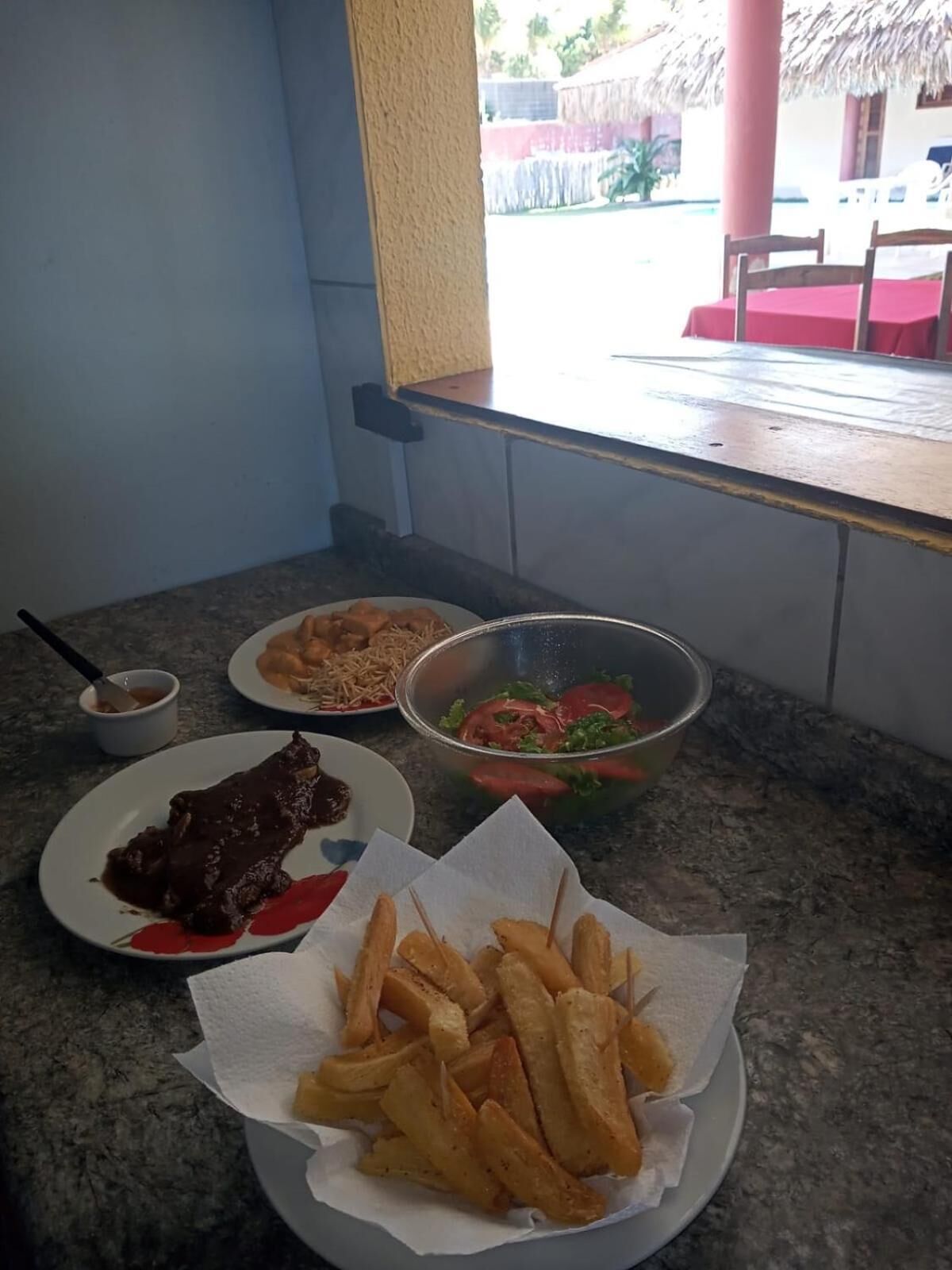 Una mesa de comedor con platos de comida, incluyendo un bistec, papas fritas y una ensalada, en una habitación con ventana y vistas al exterior.