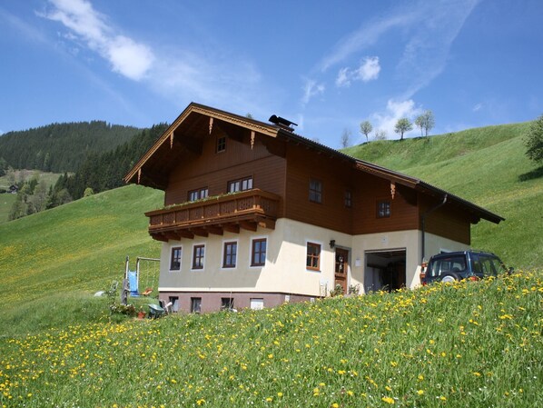 A two-story wooden house with a balcony, set in a green field with yellow flowers, under a clear blue sky.