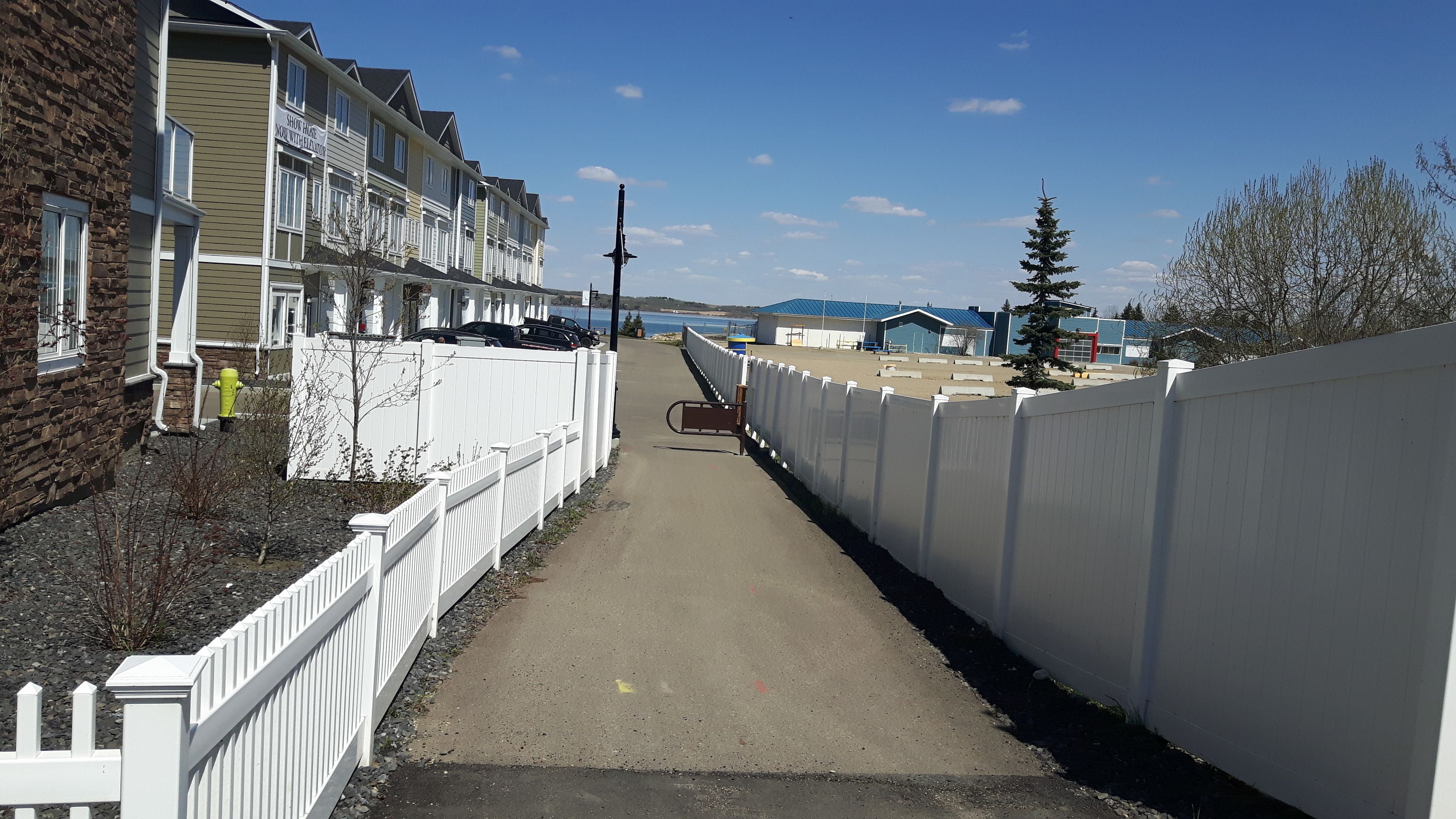 A narrow pathway flanked by white fences, leading to a row of multi-story buildings with balconies.