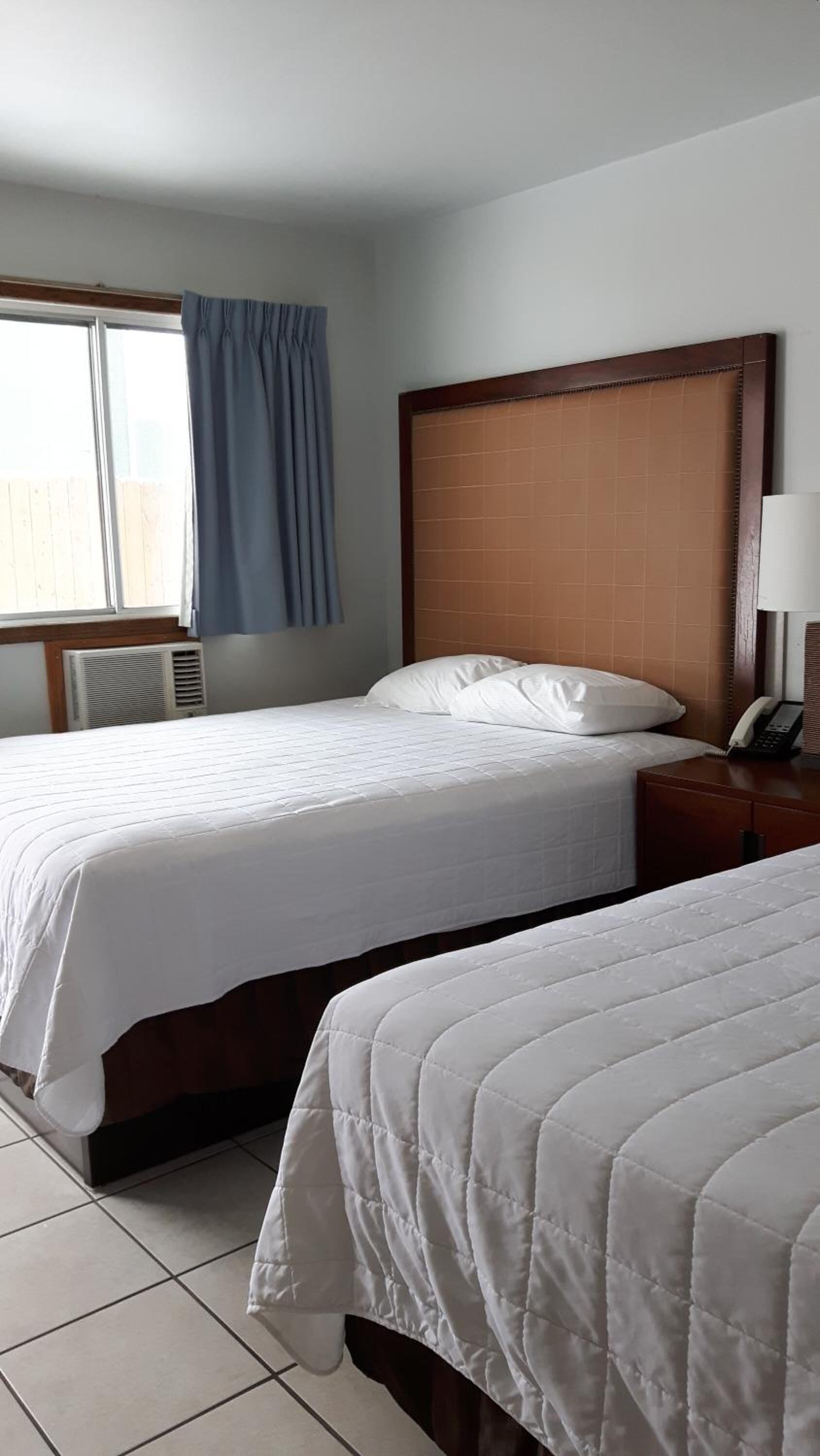 A hotel room with two beds, a wooden headboard, a window with curtains, a bedside table with a lamp, and a phone.