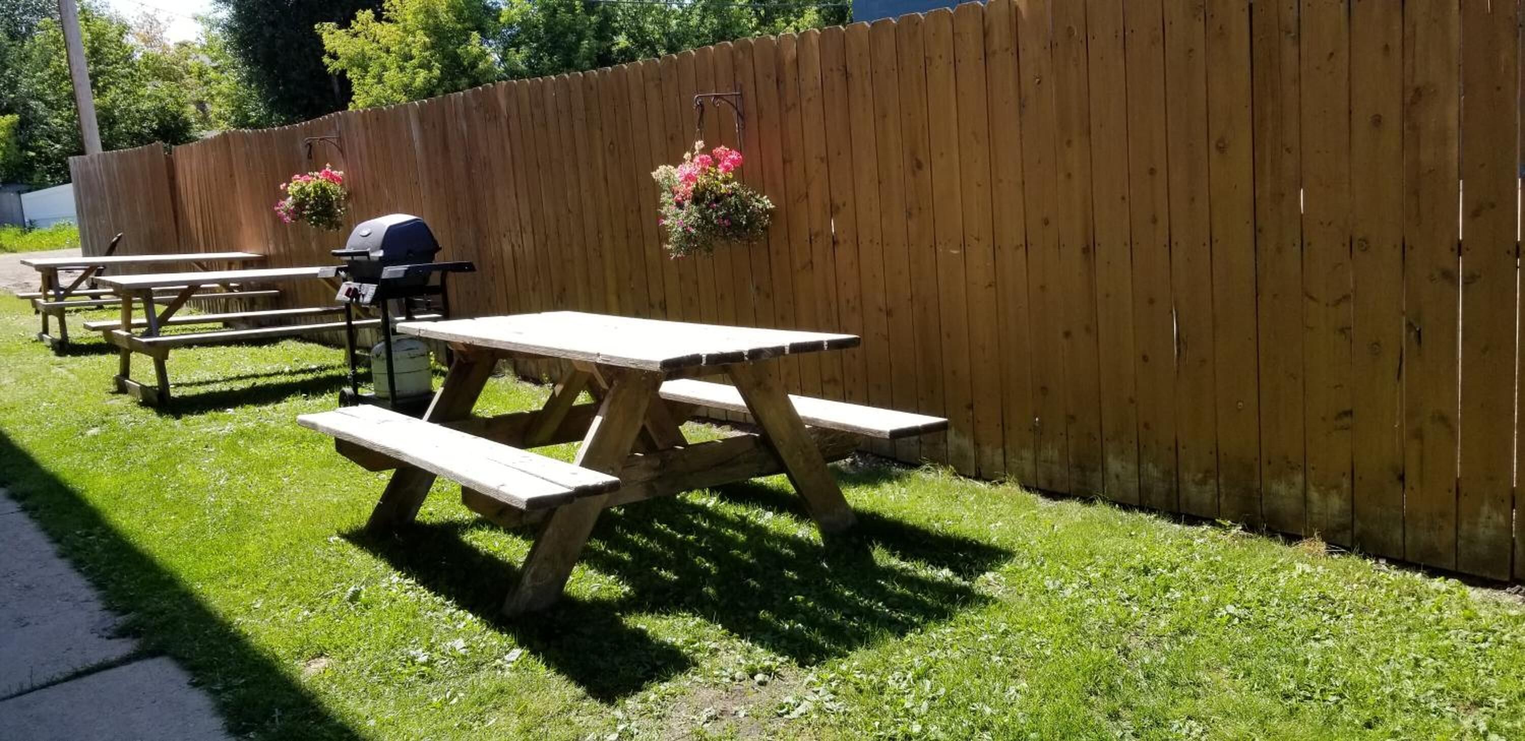 A picnic area with wooden tables and benches, a barbecue grill, and hanging flower baskets.