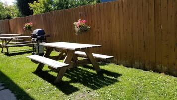 A picnic area with wooden tables and benches, a barbecue grill, and hanging flower baskets.