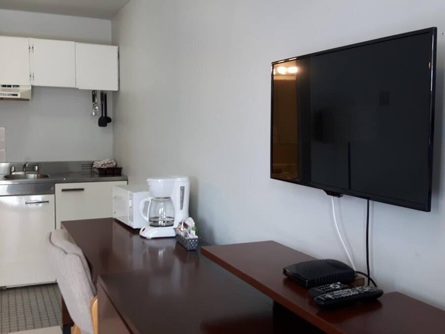 Une cuisine compacte avec des meubles blancs, une cuisinière et un four à micro-ondes. Un téléviseur à écran plat est fixé au mur, et une télécommande se trouve sur une table en bois.