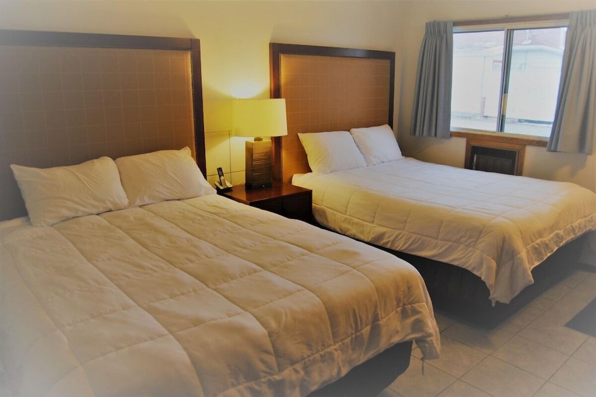 Quarto de hotel com duas camas, uma mesa de cabeceira com abajur e uma janela com cortinas.