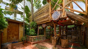 A rustic wooden structure with a thatched roof, surrounded by lush greenery and a brick-paved area.