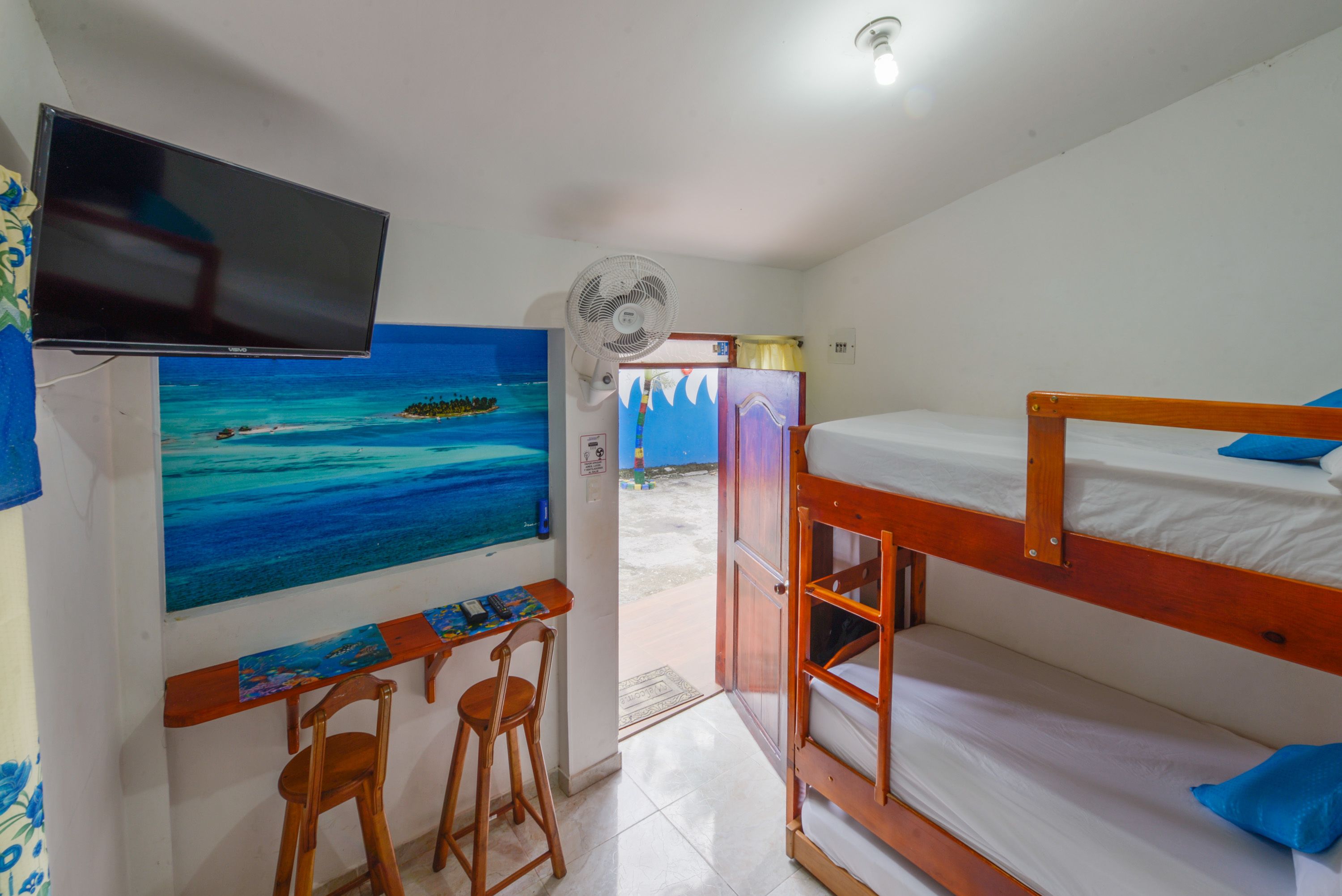 Una habitación con una cama litera, un escritorio con dos sillas, un televisor montado en la pared y un cuadro de una playa.