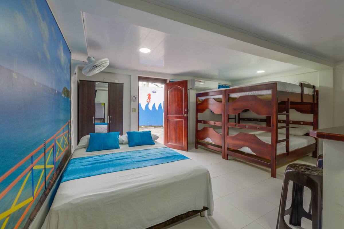 A room with bunk beds, a bed with blue pillows, a ceiling fan, and a door leading to another room.