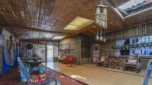 A rustic log cabin interior with a wooden floor, rocking chairs, a dining area, and a large window with decorative curtains.