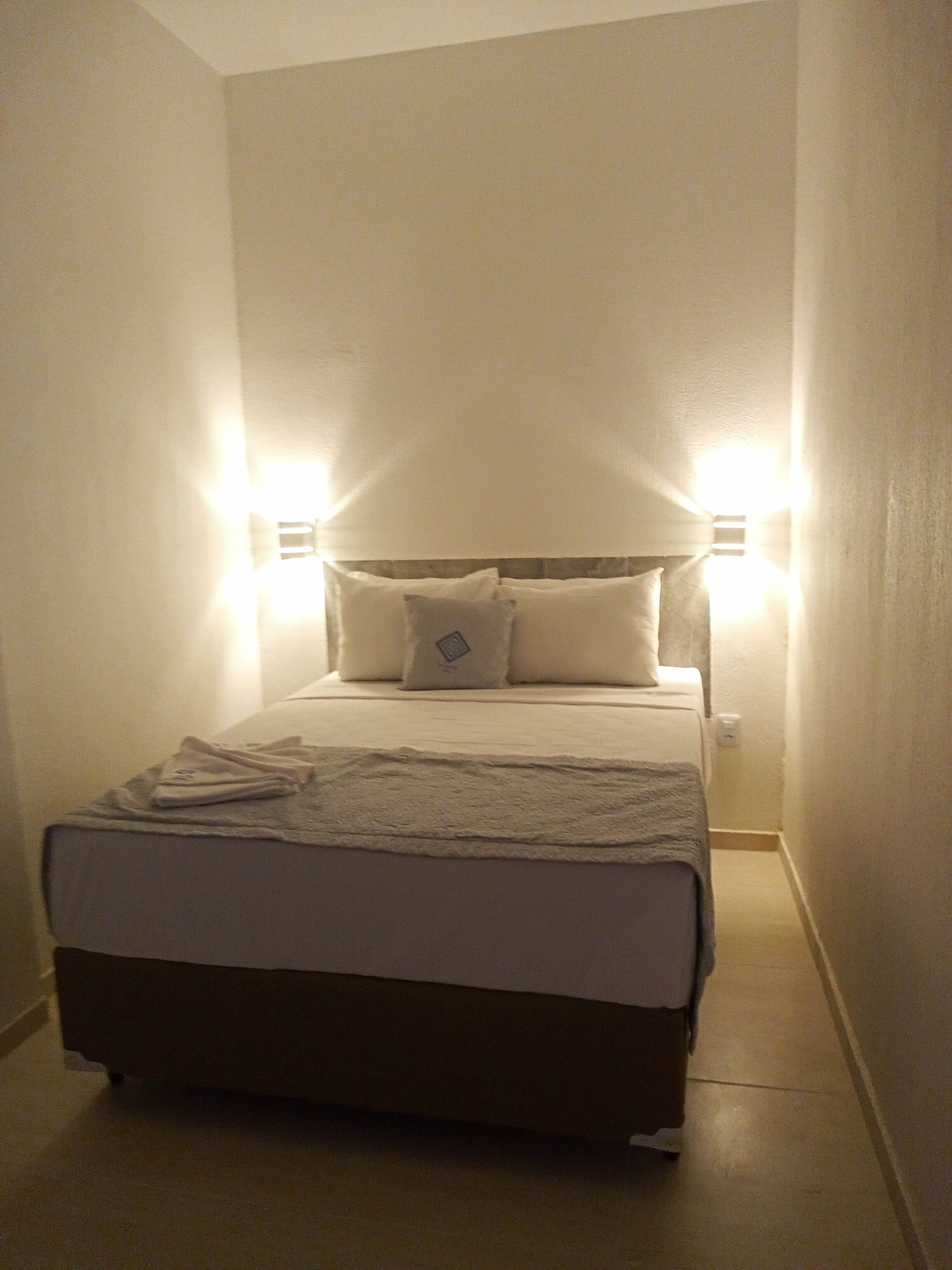 Une petite chambre d’hôtel étroite, avec un lit simple, deux lampes de chevet et une table de chevet.