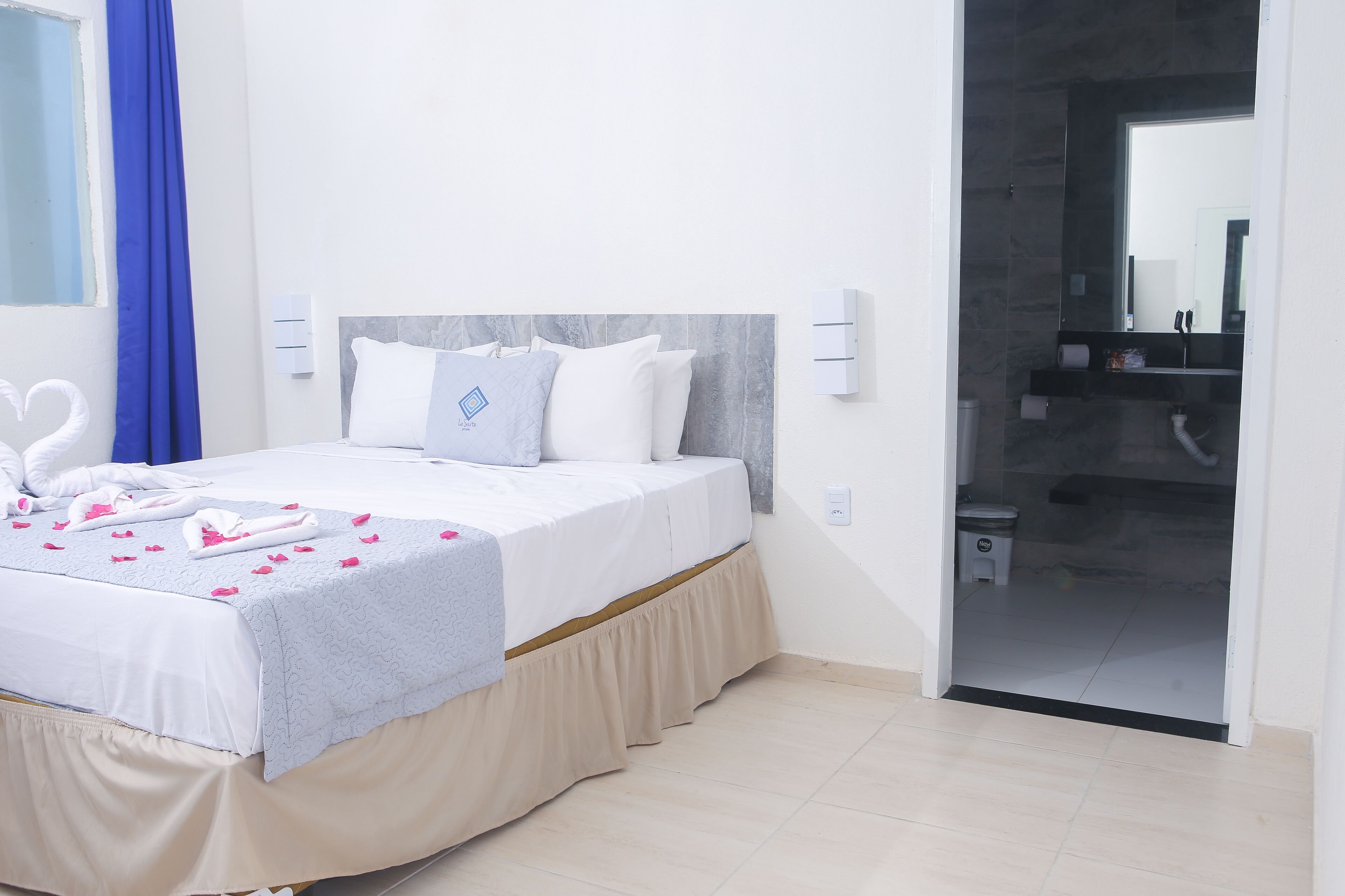 A neatly made bed with a white and light blue bedspread, a beige skirt, and pink flower petals. There is a blue curtain on the left, a white wall with a window, and a bathroom visible through an open door.
