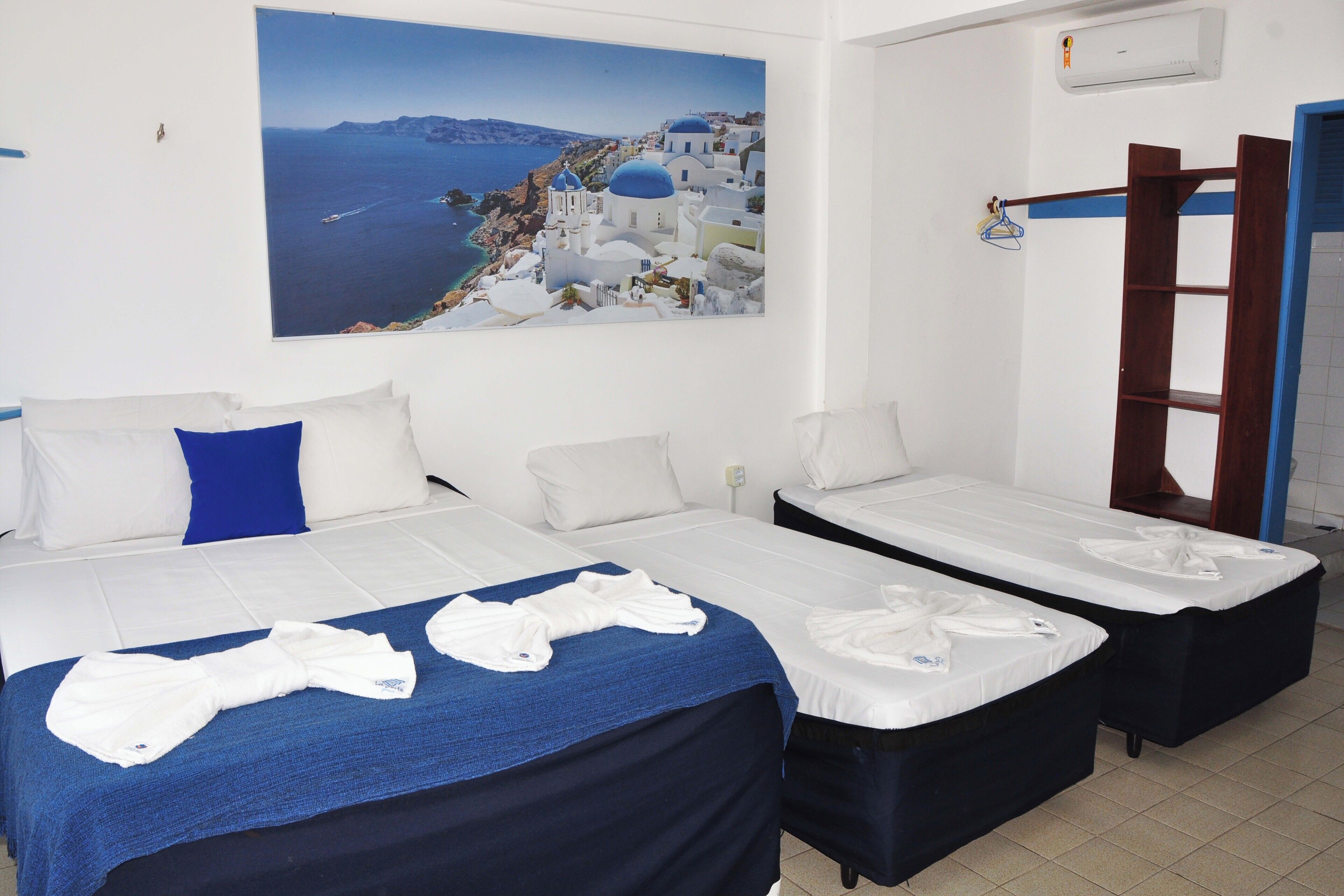 A hotel room with two beds, a blue towel on the left bed, and a painting of a coastal town on the wall.