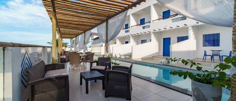 Una terraza en la azotea con mobiliario de mimbre, una piscina y un edificio con ventanas azules.