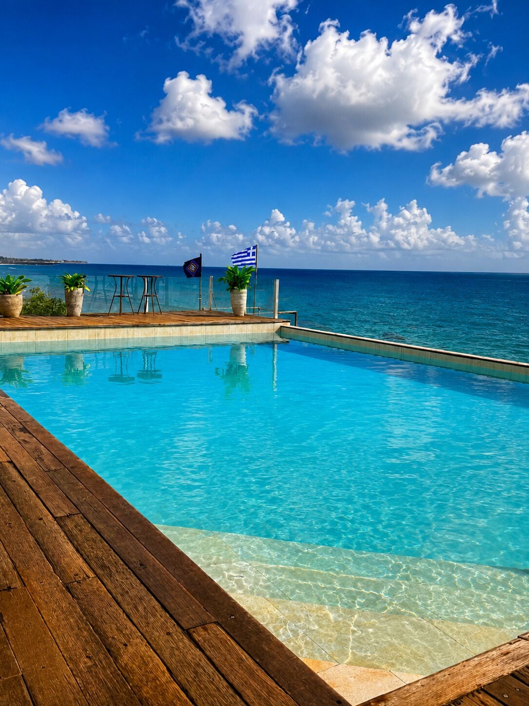 Piscina azul cristalina con muelle de madera, plantas en macetas y banderas, con vistas al mar tranquilo bajo un cielo parcialmente nublado.
