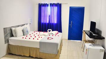 A neatly made bed with a heart-shaped decoration, a desk with a computer, and a blue door in a room with white walls.