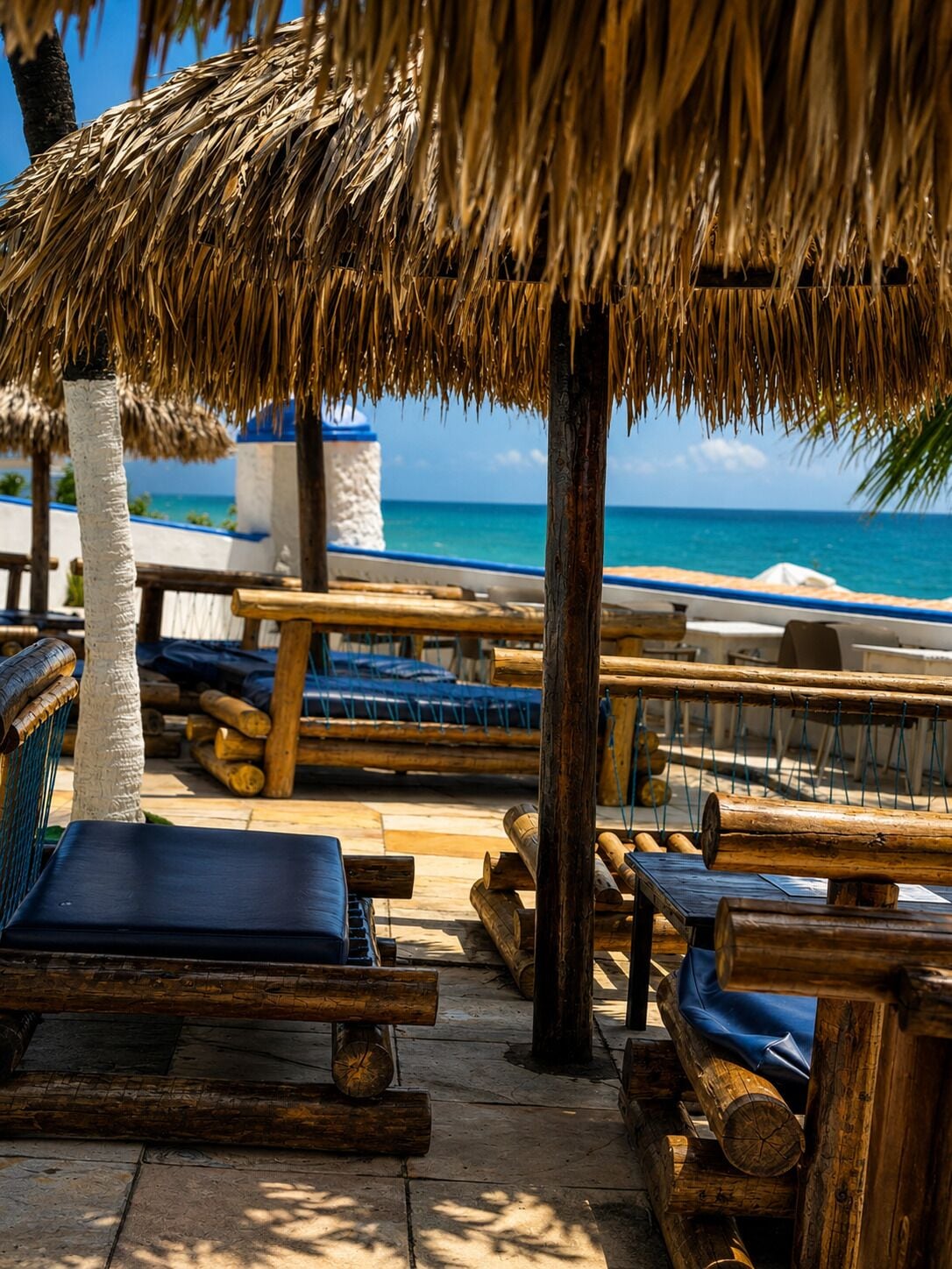 Een terras aan het strand met rieten parasols, houten ligstoelen en uitzicht op de oceaan.