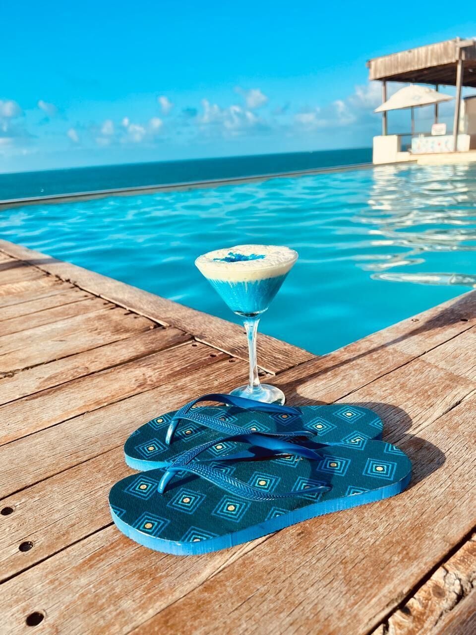 Una terraza de madera con piscina, un vaso de cóctel azul y chanclas azules.