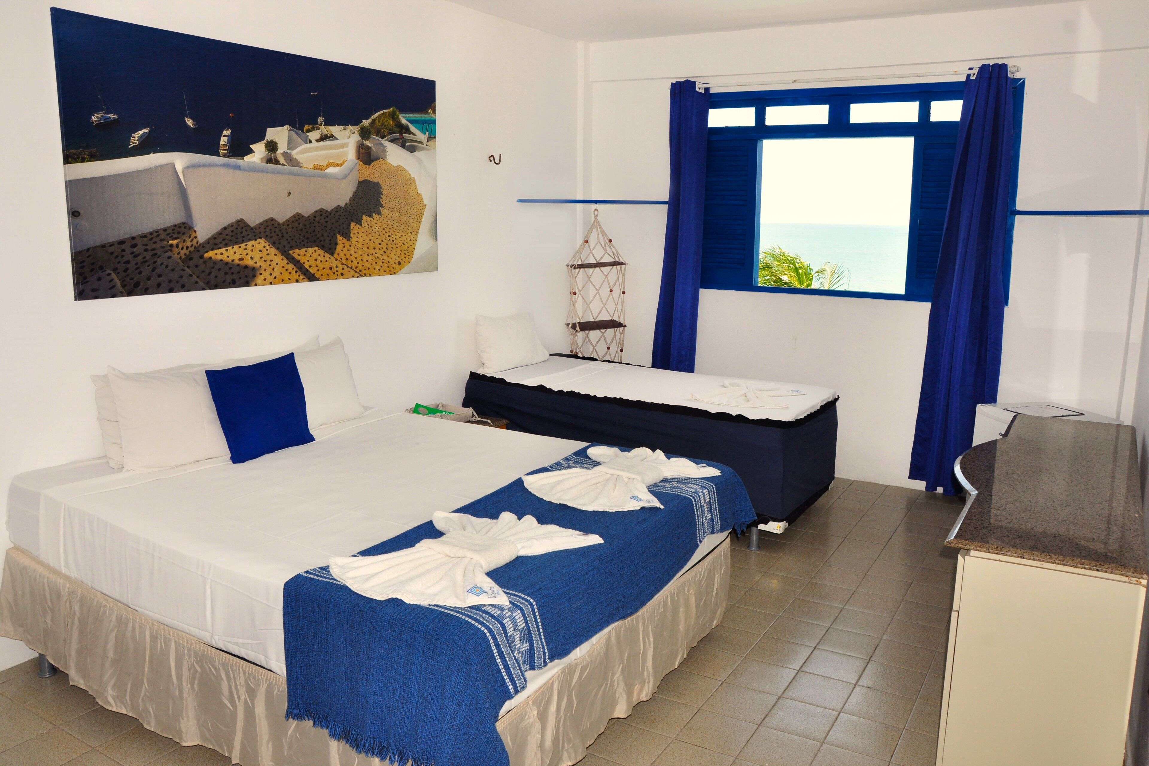 Quarto de hotel com cama, escrivaninha, cadeira, janela com cortinas e um quadro na parede.