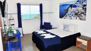 A bedroom with a bed, a desk, a chair, a small table, a plant, and a painting of a coastal scene. - La Suite Praia Hotel (Caucaia)