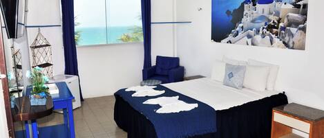 Un dormitorio con una cama, un escritorio, una silla, una mesita, una planta y un cuadro de una escena costera.