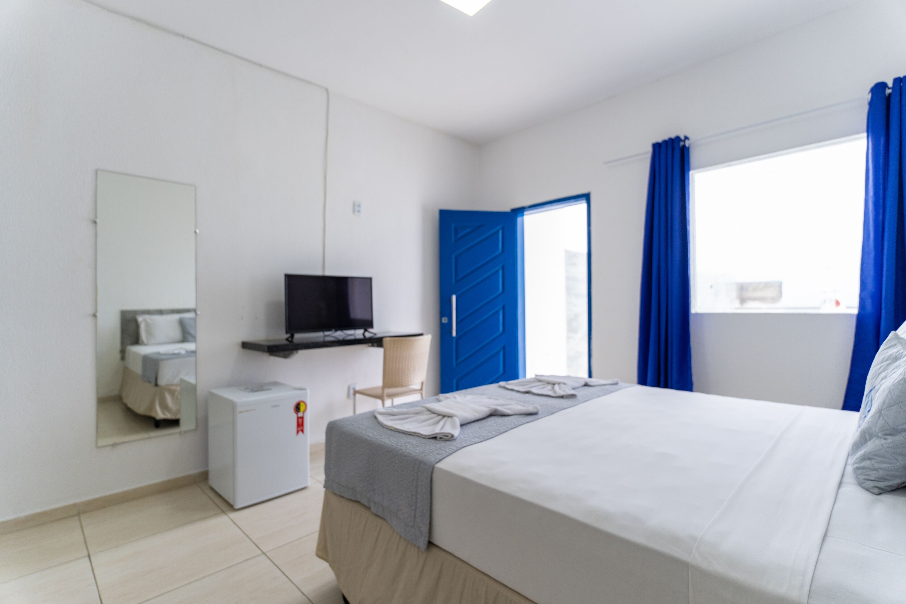 Habitación de hotel con cama, televisor, refrigerador pequeño, espejo y puerta azul.