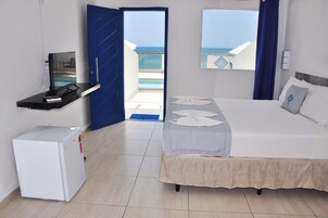 Habitación de hotel con cama, un escritorio pequeño con televisor, un refrigerador y balcón con vista al mar.