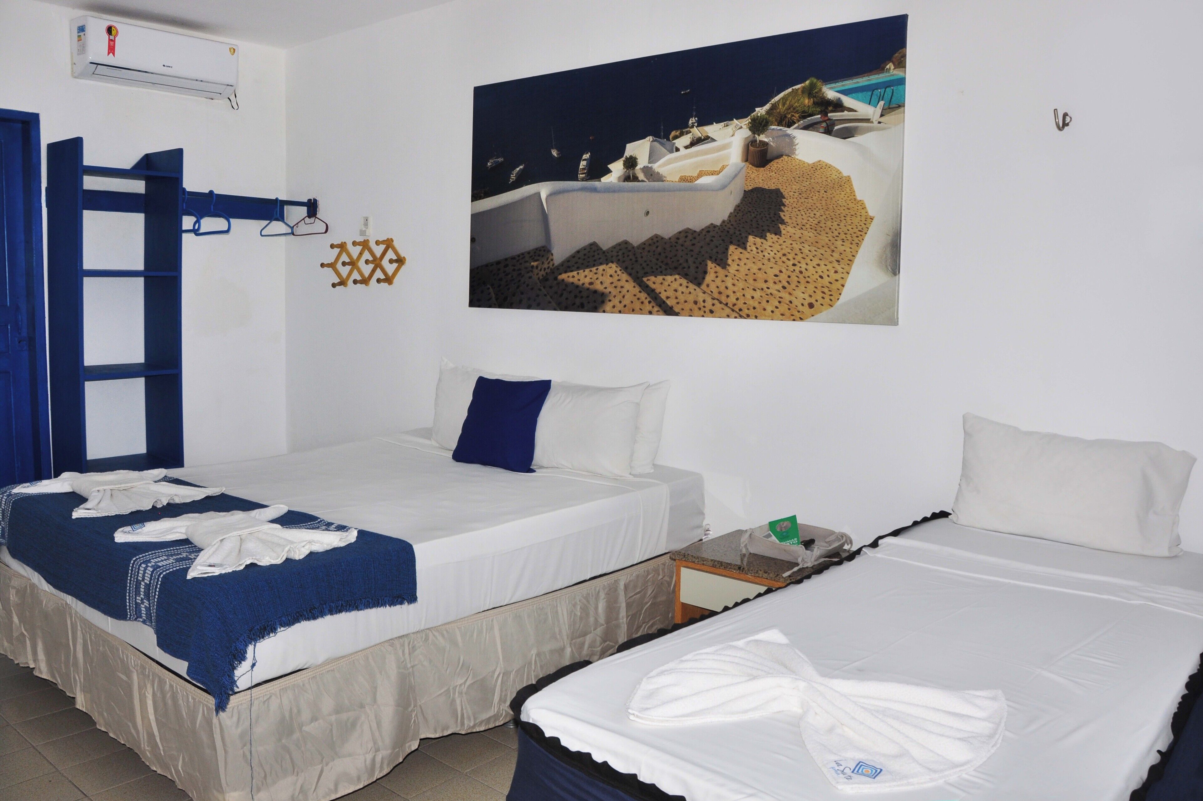 A hotel room with two beds, a blue door, a white air conditioning unit, and a painting of a coastal scene.