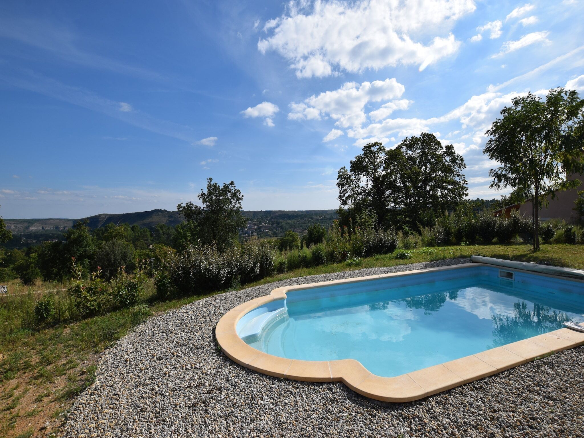 A swimming pool with a pebble border, surrounded by a gravel area, with a view of hills and trees in the background.