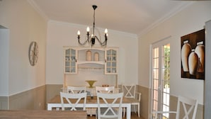 A dining area with a wooden table and white chairs, a chandelier, and a window with a view.