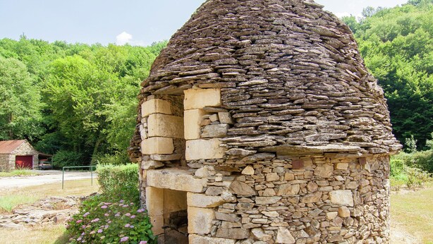 Une construction en pierre surmontée d’un toit conique, entourée de verdure, avec un petit bâtiment visible au loin.