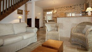 Un salon comprenant un canapé, deux fauteuils en osier, une table basse en bois, un escalier et un mur en pierre.