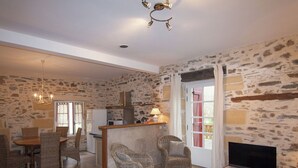 Un salon chaleureux avec un mur en pierre, un coin repas avec des meubles en osier, un téléviseur et une cuisine équipée d’un four à micro-ondes.