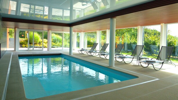 An indoor swimming pool with clear glass ceilings, sun loungers, and a view of greenery outside.