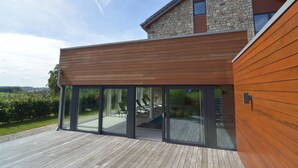 A modern house with a wooden deck, stone facade, and large sliding glass doors leading to an outdoor seating area.