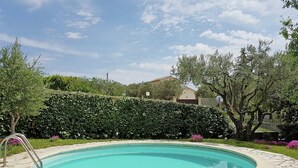 A swimming pool with a tiled deck, surrounded by a hedge and flowering plants.