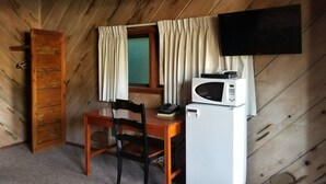Una habitación pequeña y acogedora, con techo de madera, un escritorio de madera con una silla, un refrigerador blanco, un microondas, un televisor y una ventana con cortinas.