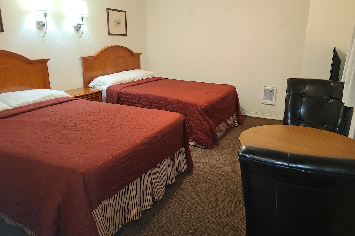 Habitación de hotel con dos camas, cabecera de madera, un sillón de cuero negro, una mesita y un respiradero en la pared.