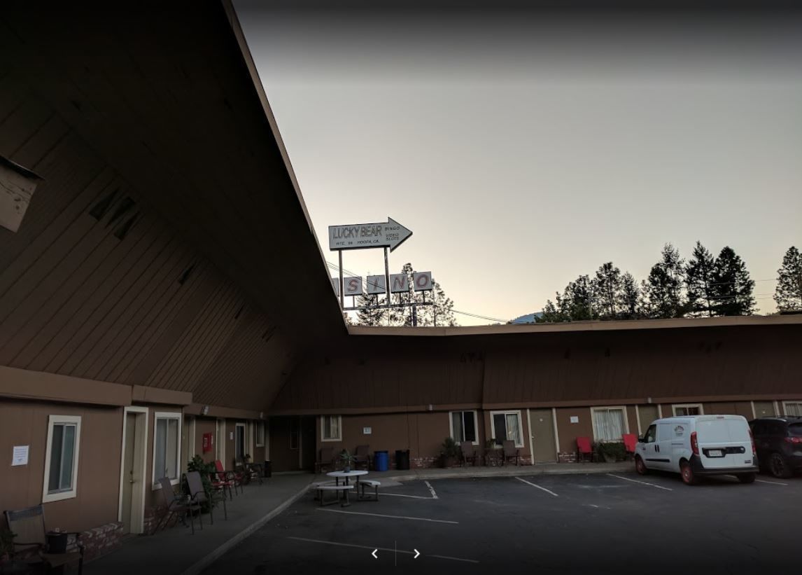 A motel with a large parking lot, a sign for 'Lucky Bear', and a row of rooms with doors and windows.