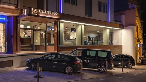 A hotel named CASTELLO with a modern facade, a glass entrance, and a parking area with two cars.