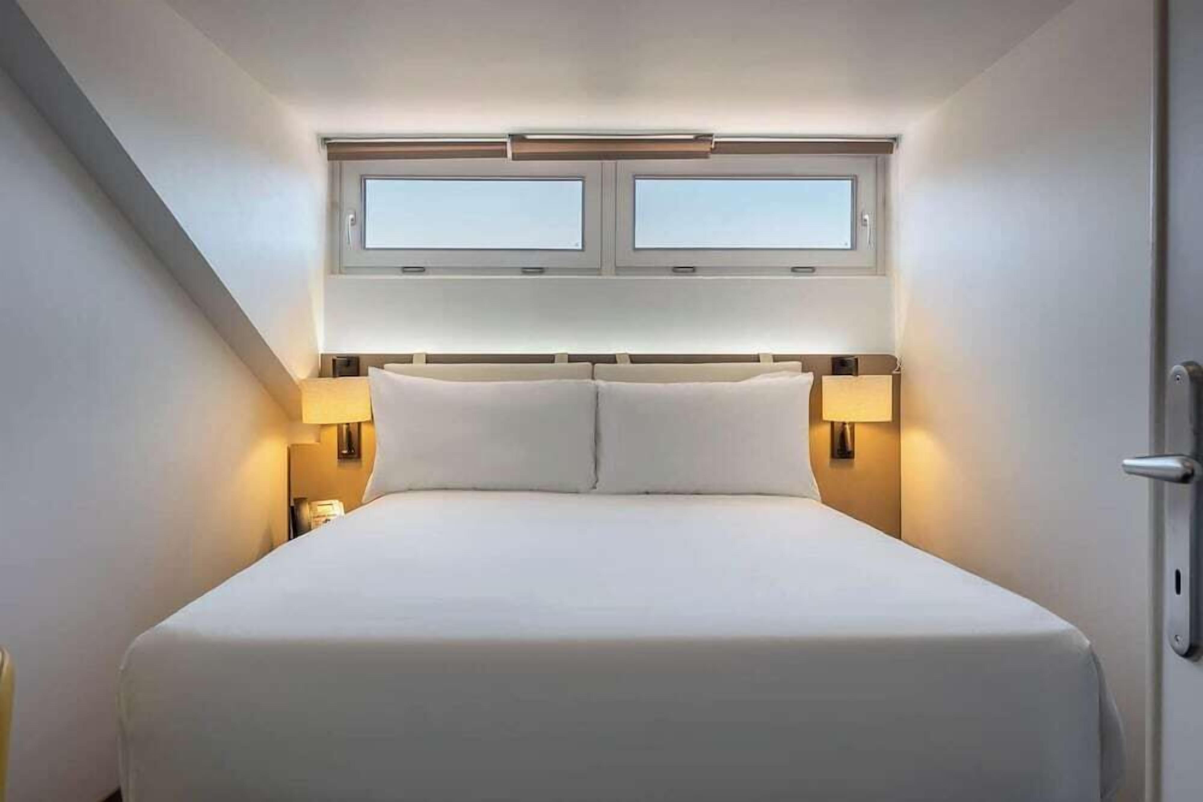 A hotel room with a large bed, two bedside lamps, a window above the bed, and a door on the right.
