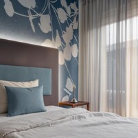Et hotelværelse med en seng, et natbord, en vase med en plante og lette gardiner.