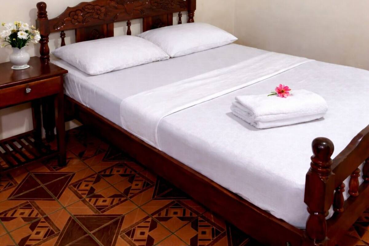 Uma cama bem arrumada com lençóis brancos, cabeceira de madeira, mesa de cabeceira com um vaso de flores e piso estampado.