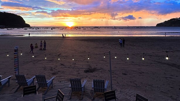 Una playa con sillas y mesas, un atardecer y barcos a lo lejos.