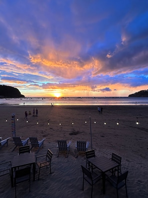 A beach with chairs and tables, a sunset, and boats in the distance.