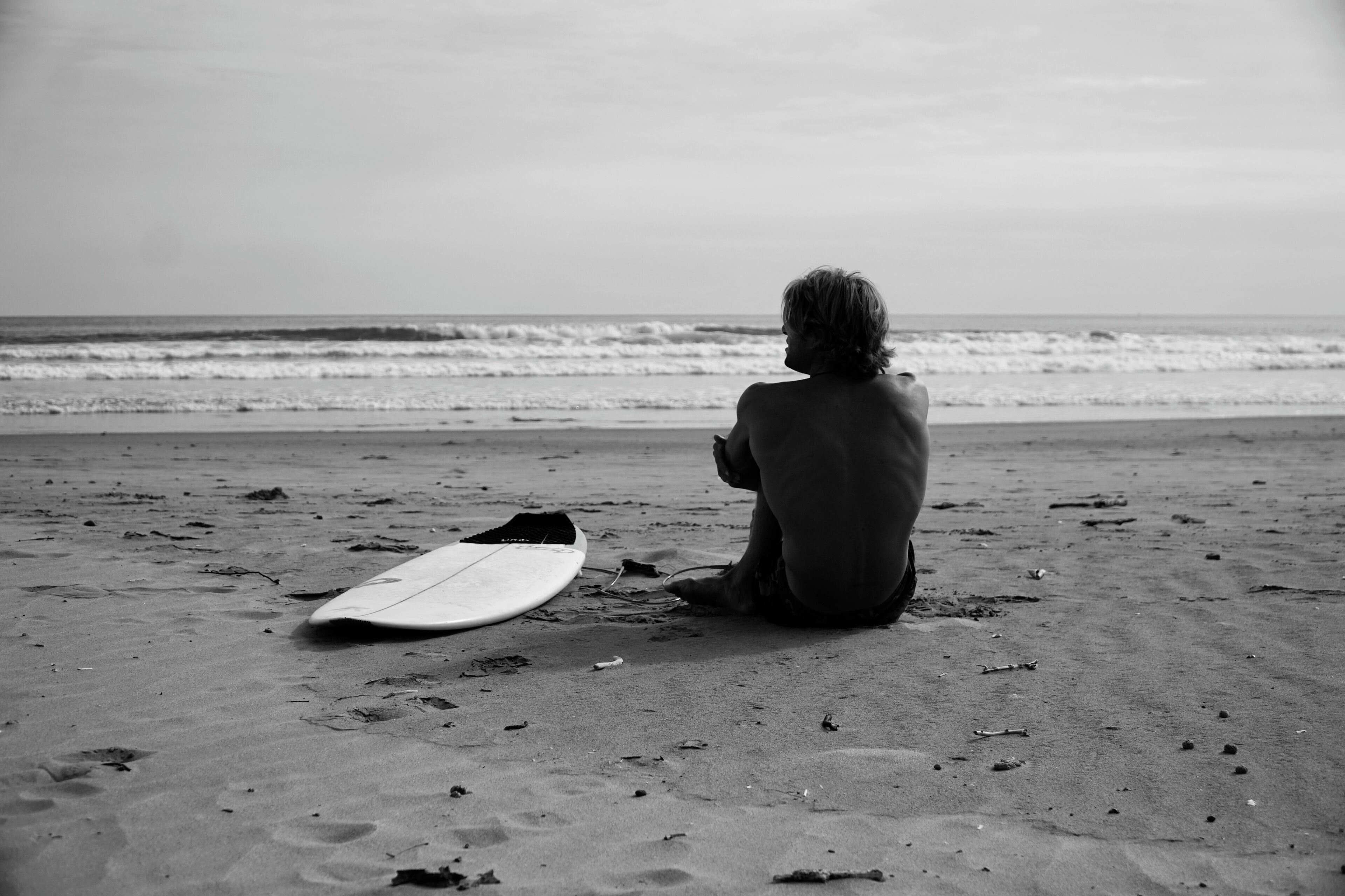 Una persona sentada en la playa con una tabla de surf a su lado.