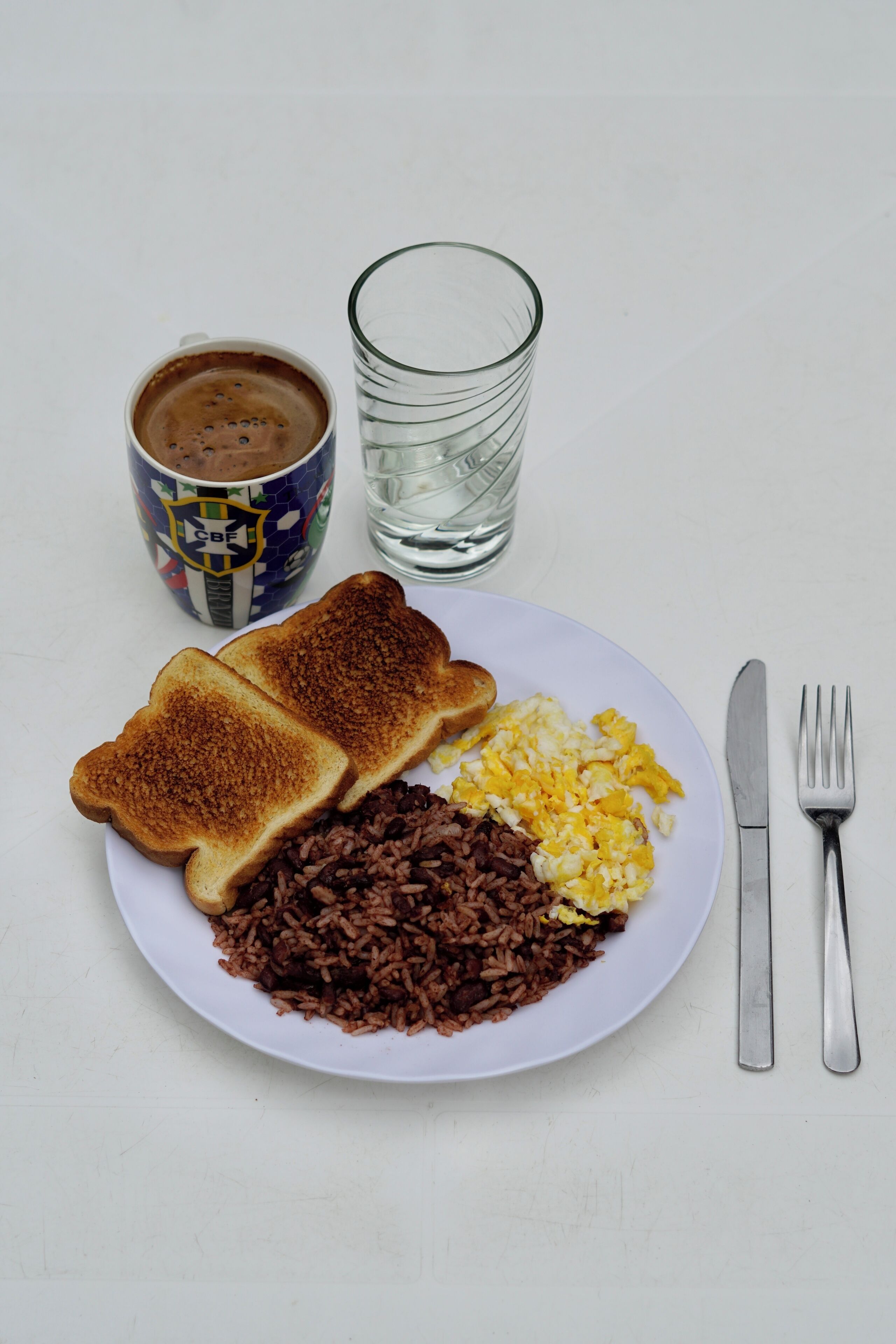 Prato com torradas, ovos mexidos e arroz integral, uma xícara de café e um copo de água sobre uma mesa branca.